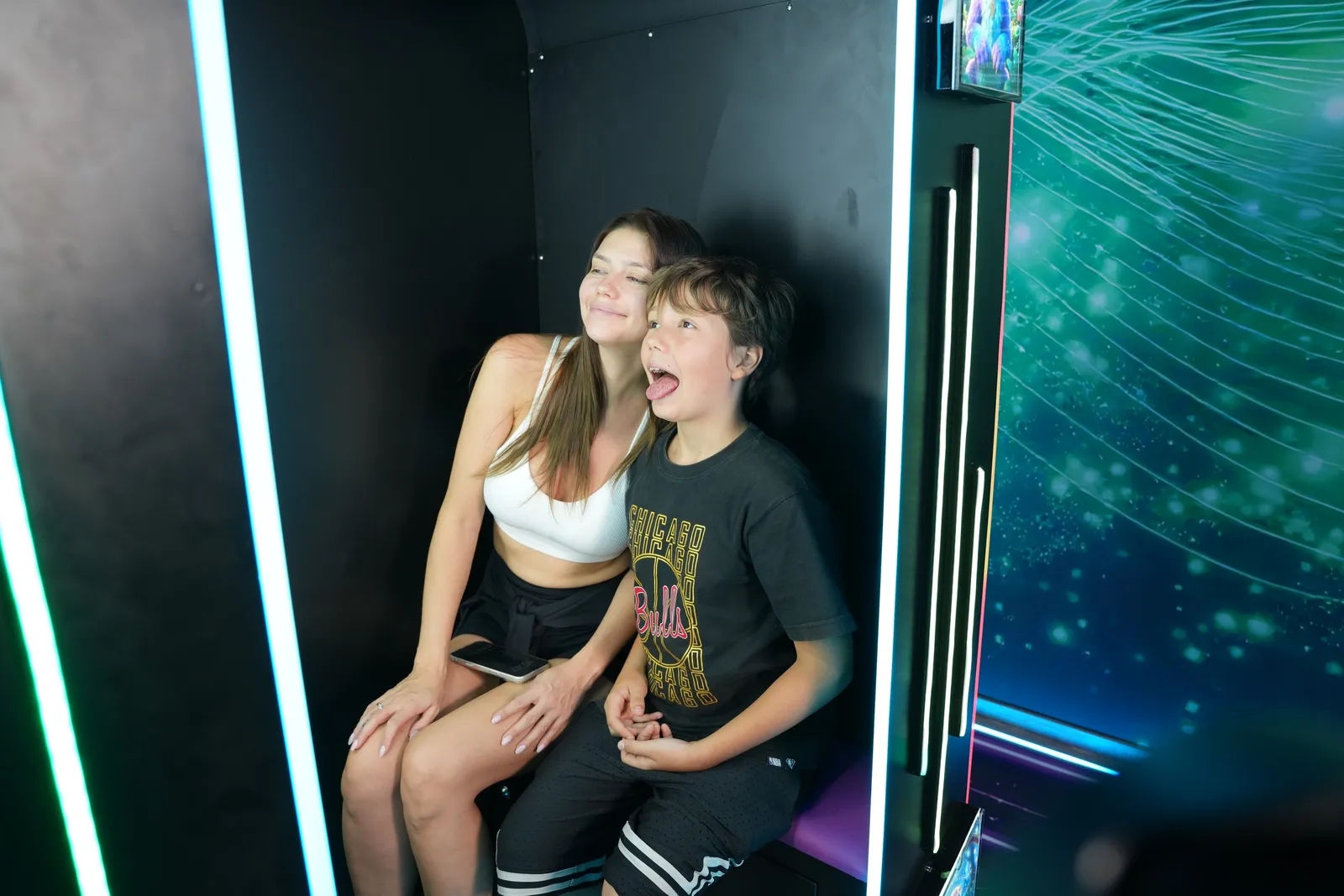 Mother and son laughing inside a Futureino AI Photo Booth vending machine lit by neon strips while the screen captures their photo